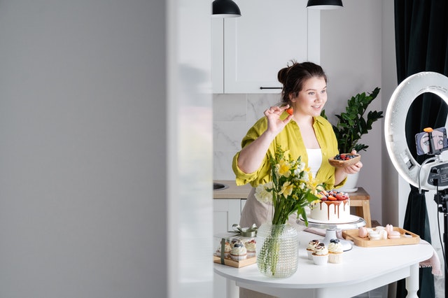 Woman holding a strawberry and a berry bowl with a yellow blouse in the kitchen smiling into a camera phone with LED ring light. 