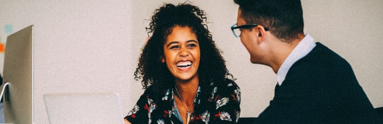 Smiling woman in blouse, sitting at laptop and talking to man in sweater and glasses.