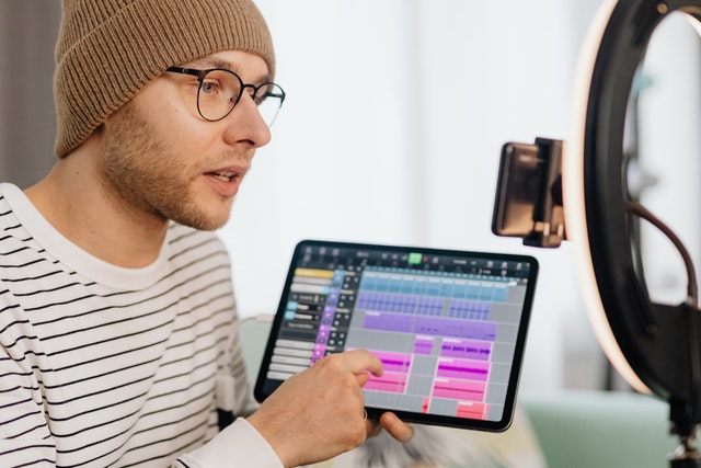 Man with beanie hat, glasses, and striped black-and-white shirt points to music app on a tablet looking into iPhone with LED ring light