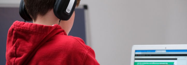 Student in red hoodie with headphones, sitting at desk in school and working on laptop.