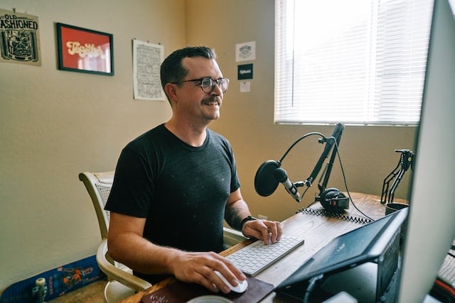 Man with mustache and glasses smiling at computer with podcast setup