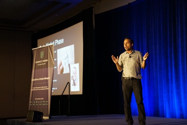 Man in casual button-up and black jeans stands on a stage with a presentation format and blue curtain behind him.