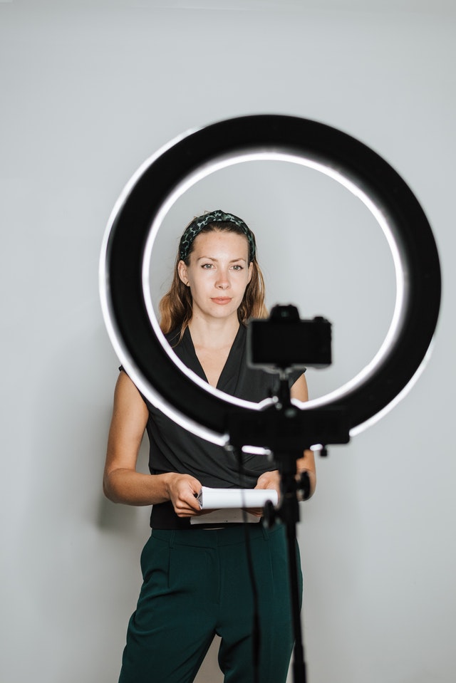 Woman in black outfit with notebook looking into camera on tripod against an LED ring light.