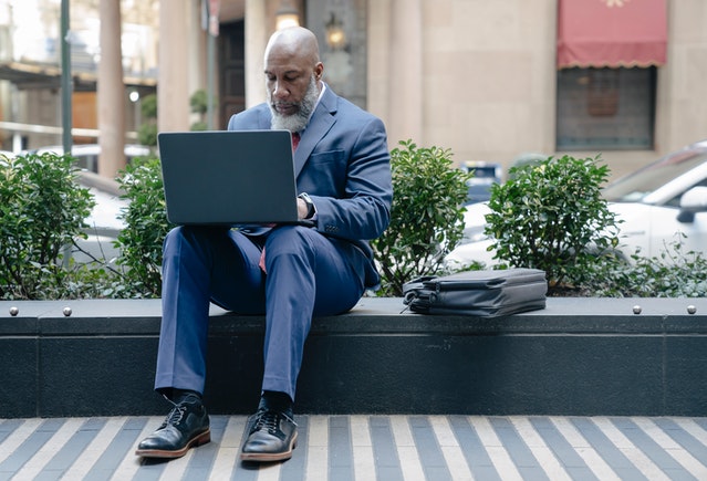 Business man in blue suit sitting outside a building on a computer.