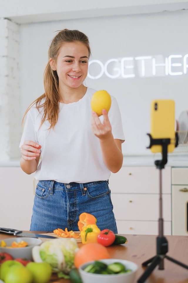 Woman in white shirt and jeans holding a lemon in the kitchen and smiling into an iPhone camera on a tripod 