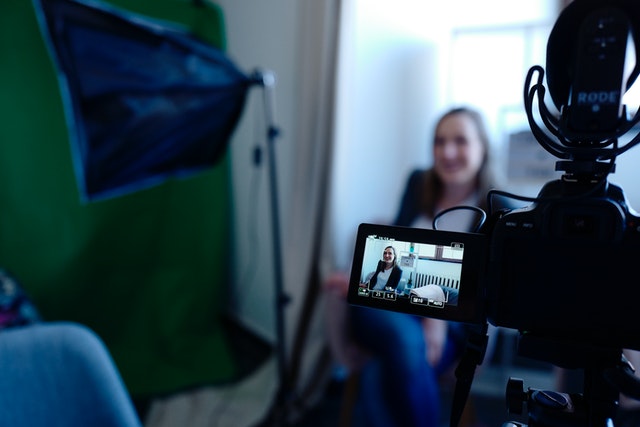 Woman sits behind a camera smiling on a set with green screen nearby