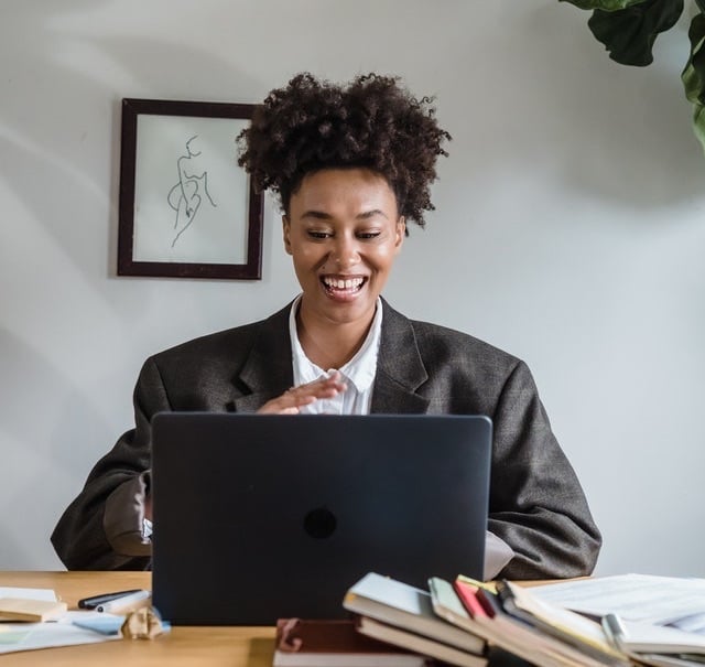 Woman_in_brown_blazer_smiling_and_gesturing_behind_a_laptop_against_white_wall_with_picture_frame_and_leafy_plant