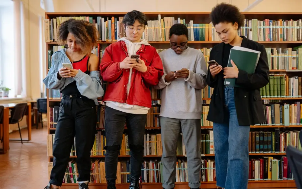 Four students preoccupied on phones looking down in front of a stack of library books.