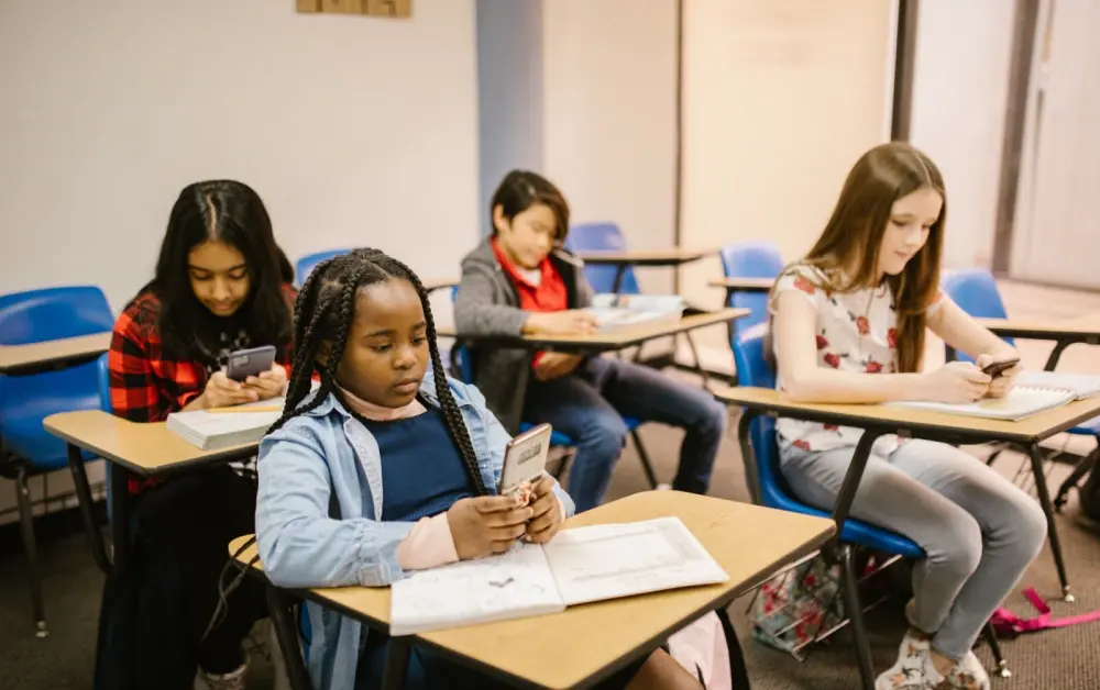 Four young learners all preoccupied on phones sitting at desks in the classroom.