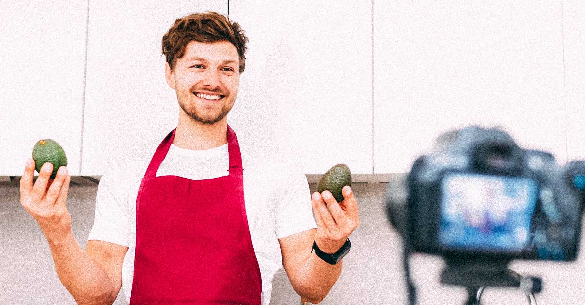 Man filming video tutorial about cooking, wearing an apron and holding an avocado up to a camera tripod