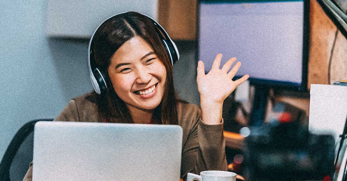 Woman smiling and waving at laptop while wearing headphones and watching to a screencast