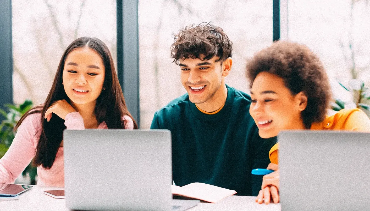 Three smiling students (one male and two females) behind laptops working together