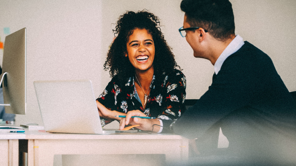 Smiling woman in blouse, sitting at laptop and talking to man in sweater and glasses.