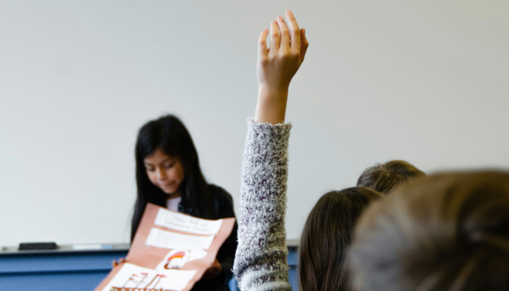 Student presenting at the front of a classroom with another student in the background raising their hand.
