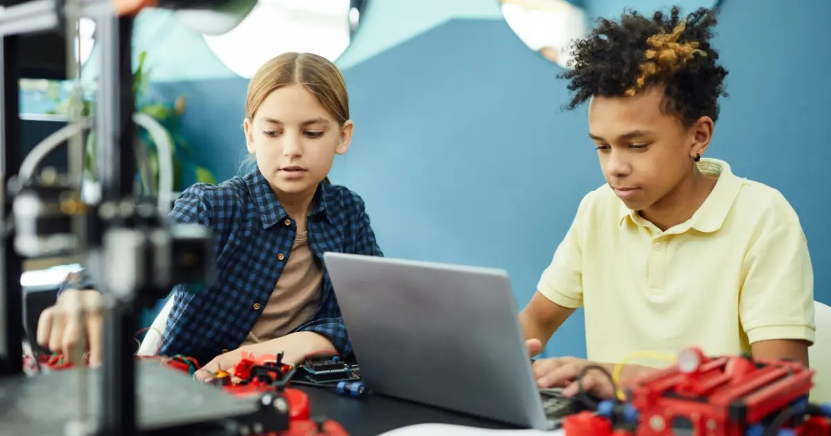 Two students sitting next to each other in science class: One interacting with objects and one typing on the laptop.