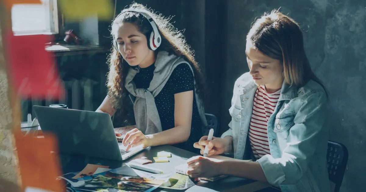 Two female students sitting next to each other: One with headphones and writing on a laptop and the other storyboarding next to her.