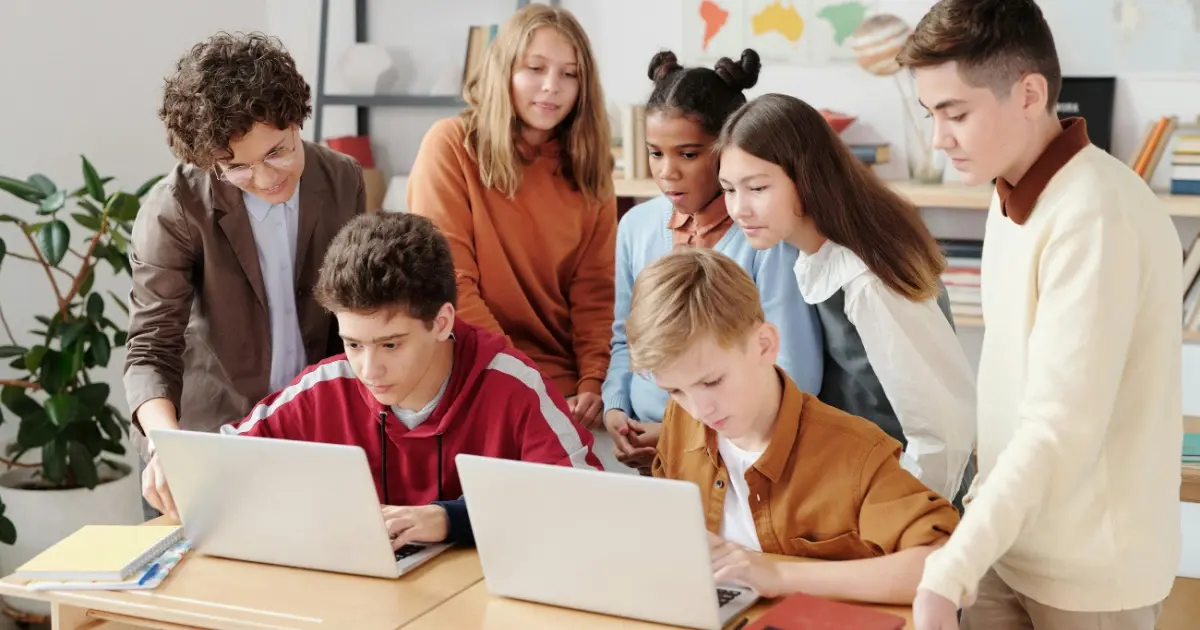 Class of students with two sitting behind laptops and peers standing behind them collaborating on a project.