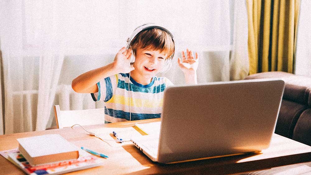 Elementary student waving to computer while wearing headphones and watching video