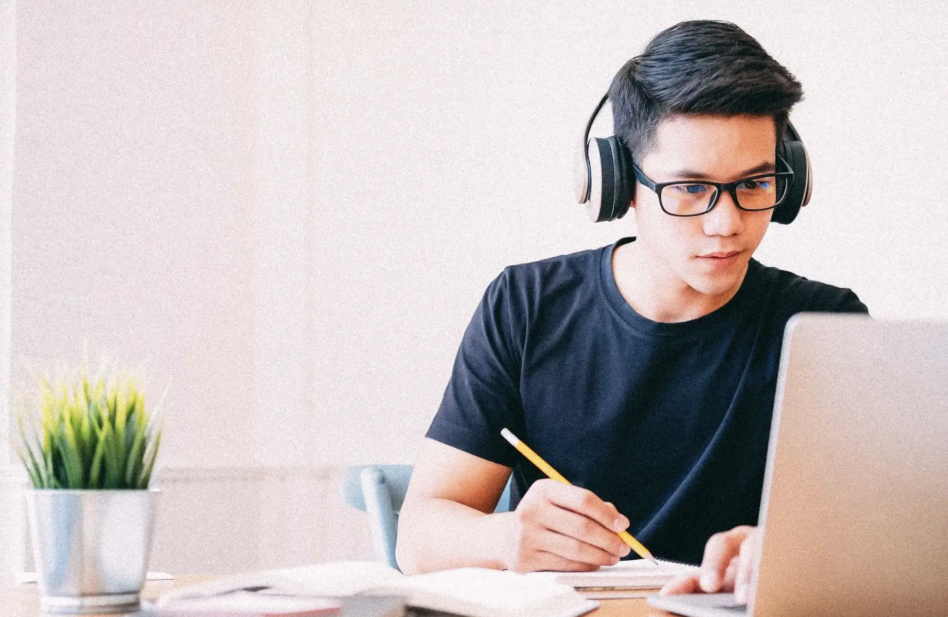 An adolescent student takes notes with a pencil while listening to a lesson on a laptop.