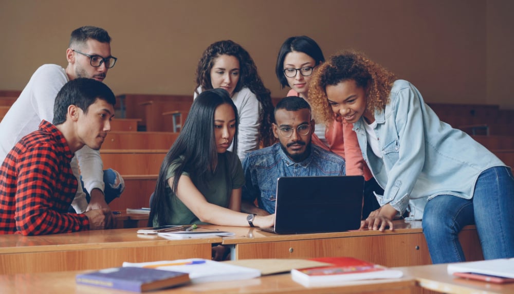 Group of high school or college age students looking over one student's laptop in a classroom setting.
