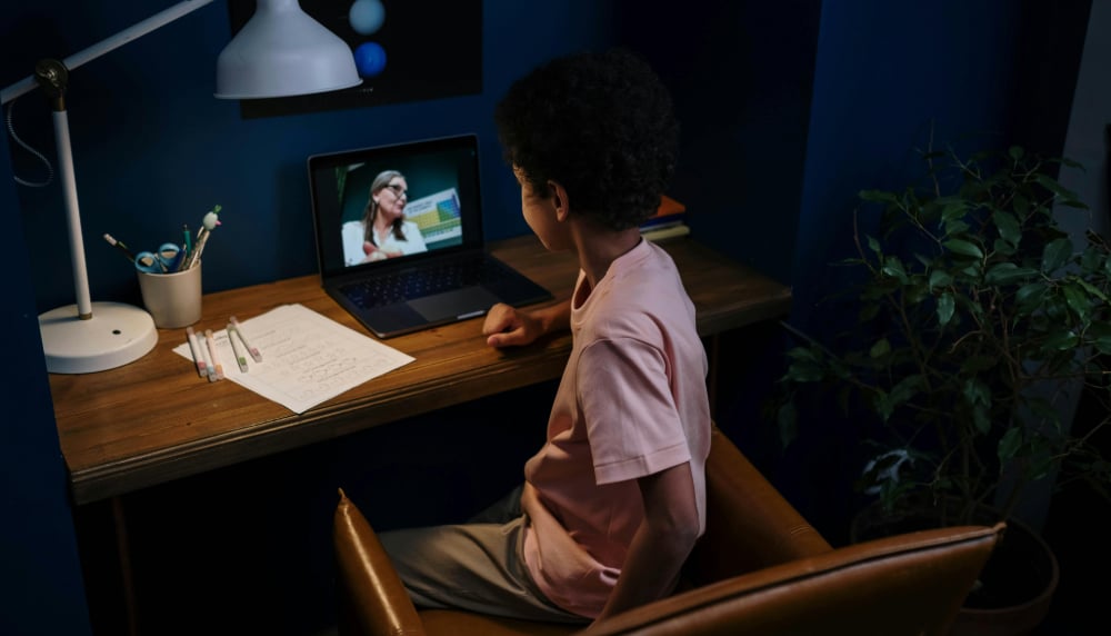 Young student looking over his laptop on his desk at home with a person on screen and notebook close by.