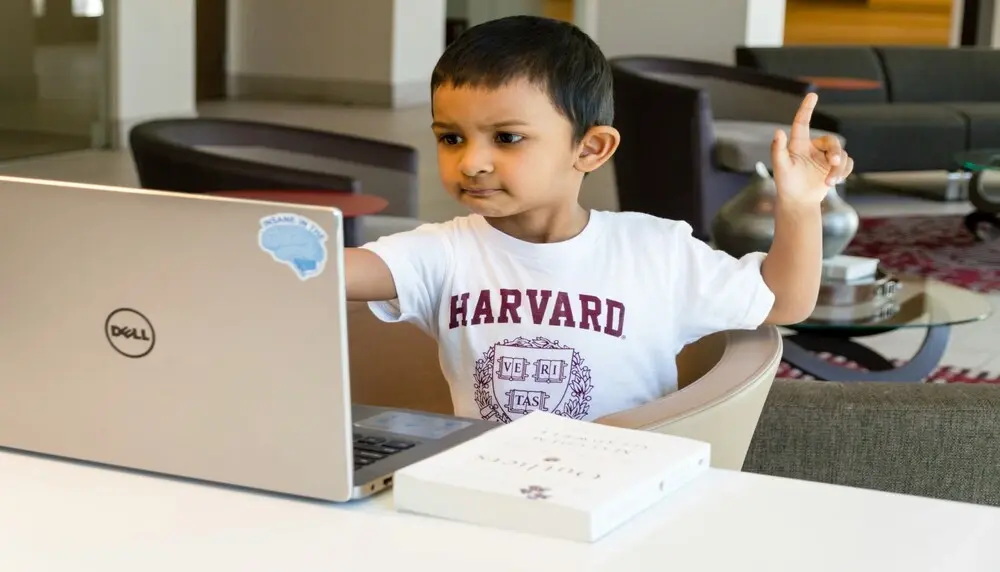 Boy in Harvard t-shirt sits in front of a computer with one finger in the air as he engages