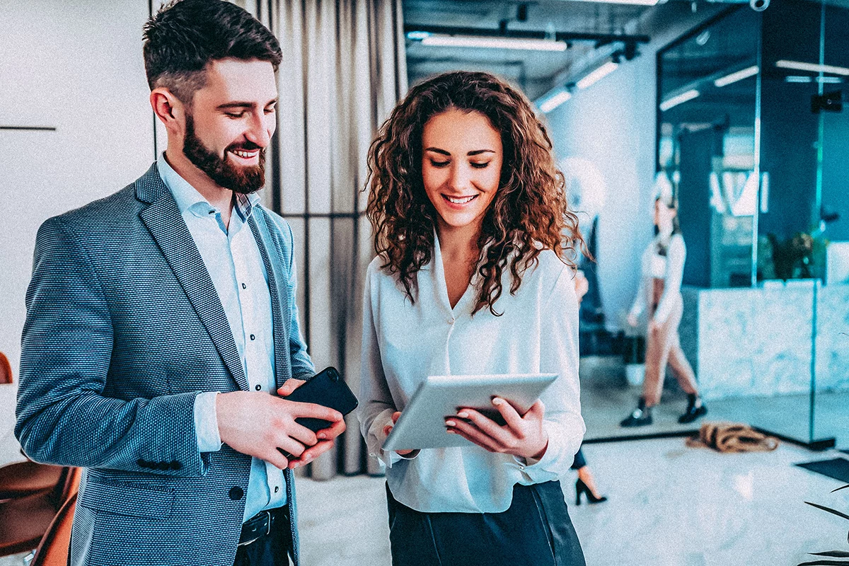 One male and one female colleague smiling while viewing video content on a tablet in an enterprise office