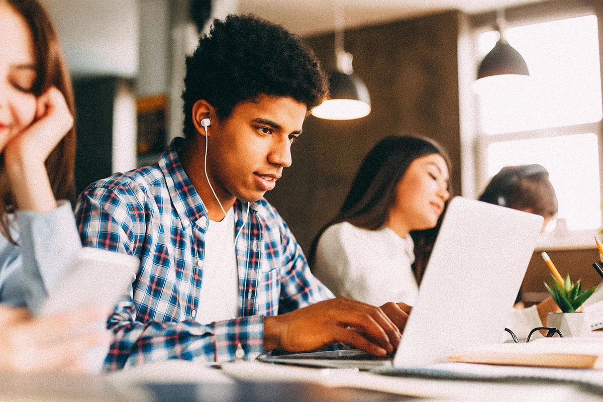 Young adult in flannel shirt working on laptop with headphones in his ears. Other young adults around him.