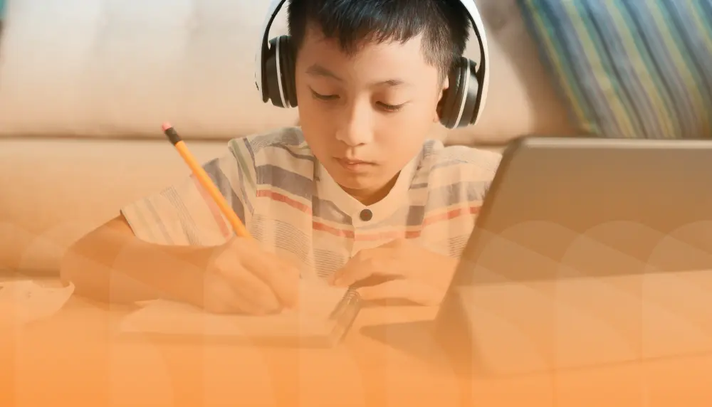 A young student taking notes with a pencil, listening to a lesson on a tablet device.