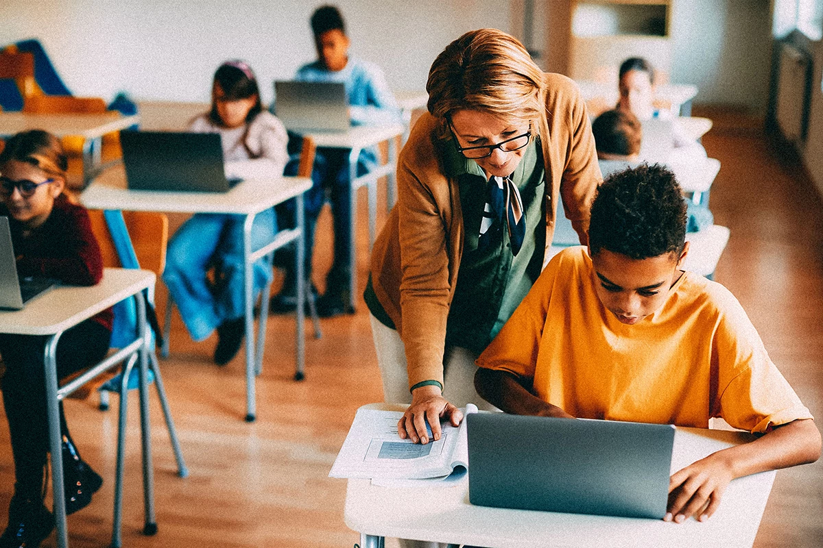 Teacher working with student on laptop in classroom. 