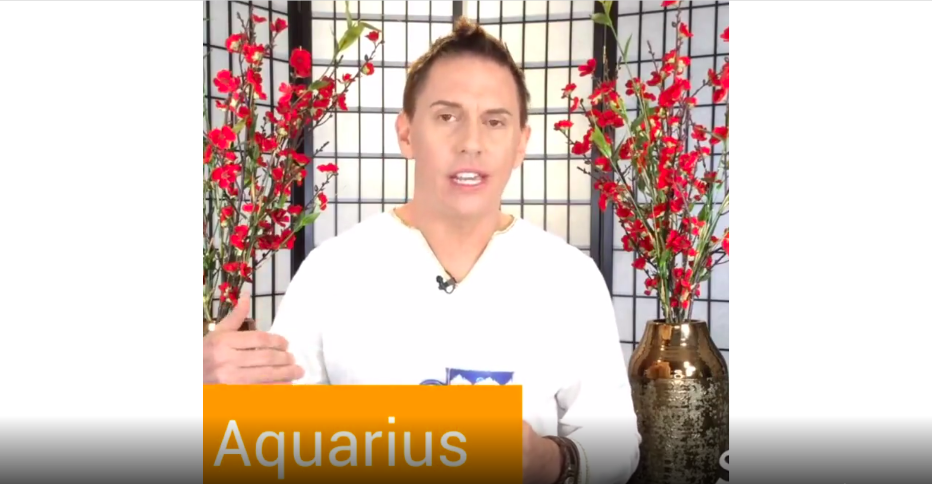 Man talking to camera in front of Asian paper screen and red flowers with word AQUARIUS as title card