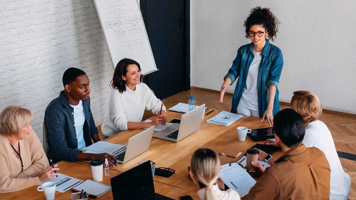 Group of young employees at meeting table. One woman is presenting to the group.