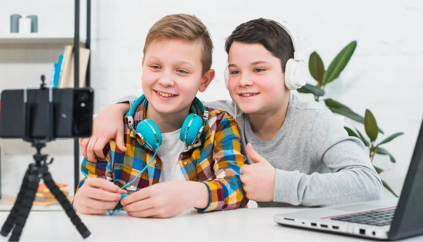Two boys smiling at a camera with a laptop and headphones as they record a video for school communications