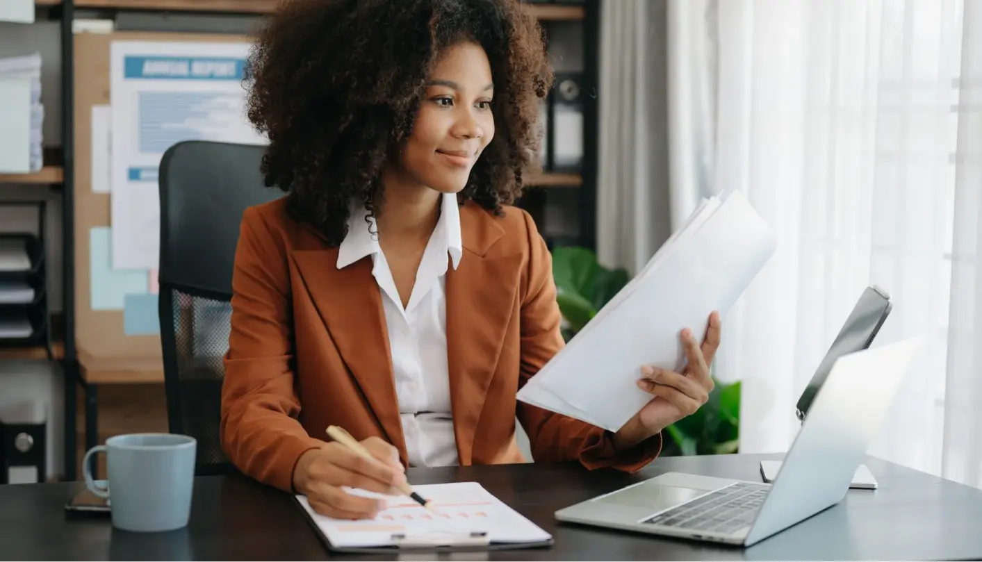 Confident businesswoman reviews a document and takes notes on a pad in front of her laptop.