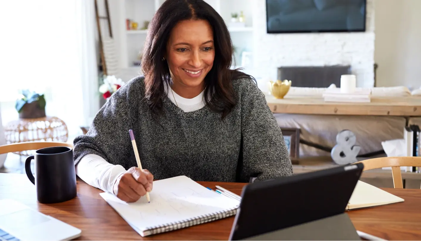 Smiling woman sits at a table taking notes while engaging with a tablet device.