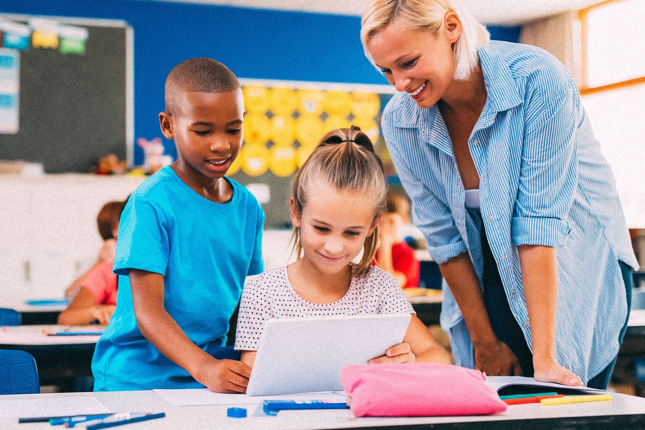 Two elementary-aged children and their teacher, looking at something on an iPad.