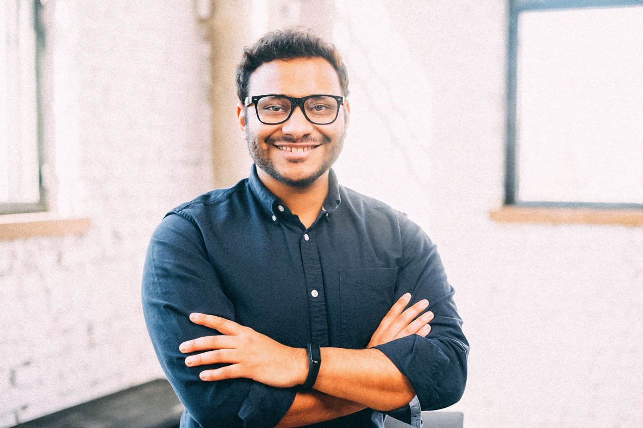 Man with glasses and dark blue collared shirt, in open area room, facing camera with arms folded and smile on face.